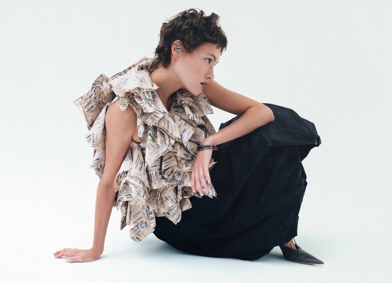 Model with short dark hair wearing a patterned paper ruffle top and black skirt seated against a white background.