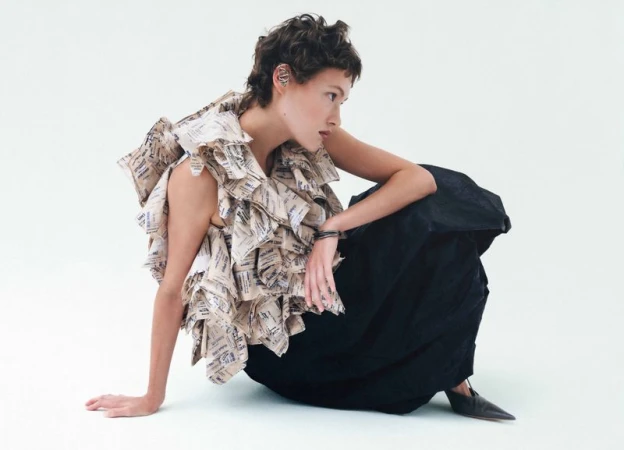 Model with short dark hair wearing a patterned paper ruffle top and black skirt seated against a white background.
