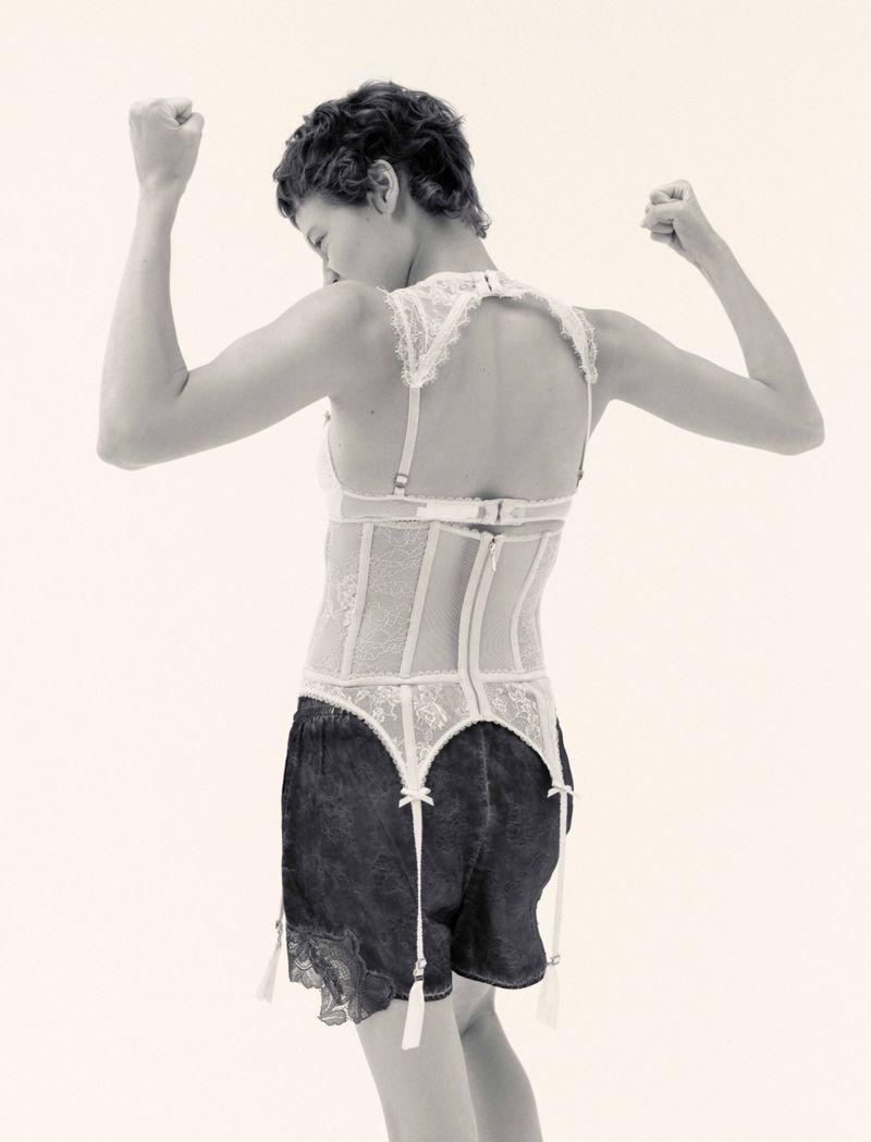 Model with short dark hair wearing a white lace corset and black shorts posing against a light background