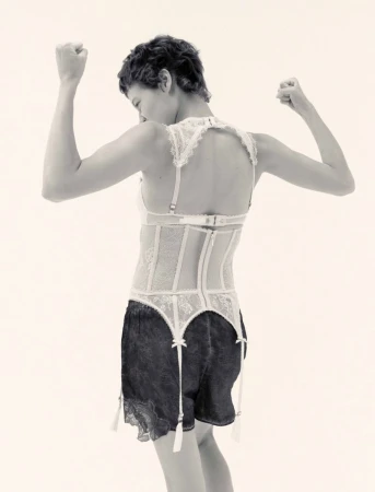 Model with short dark hair wearing a white lace corset and black shorts posing against a light background