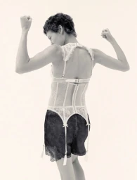 Model with short dark hair wearing a white lace corset and black shorts posing against a light background