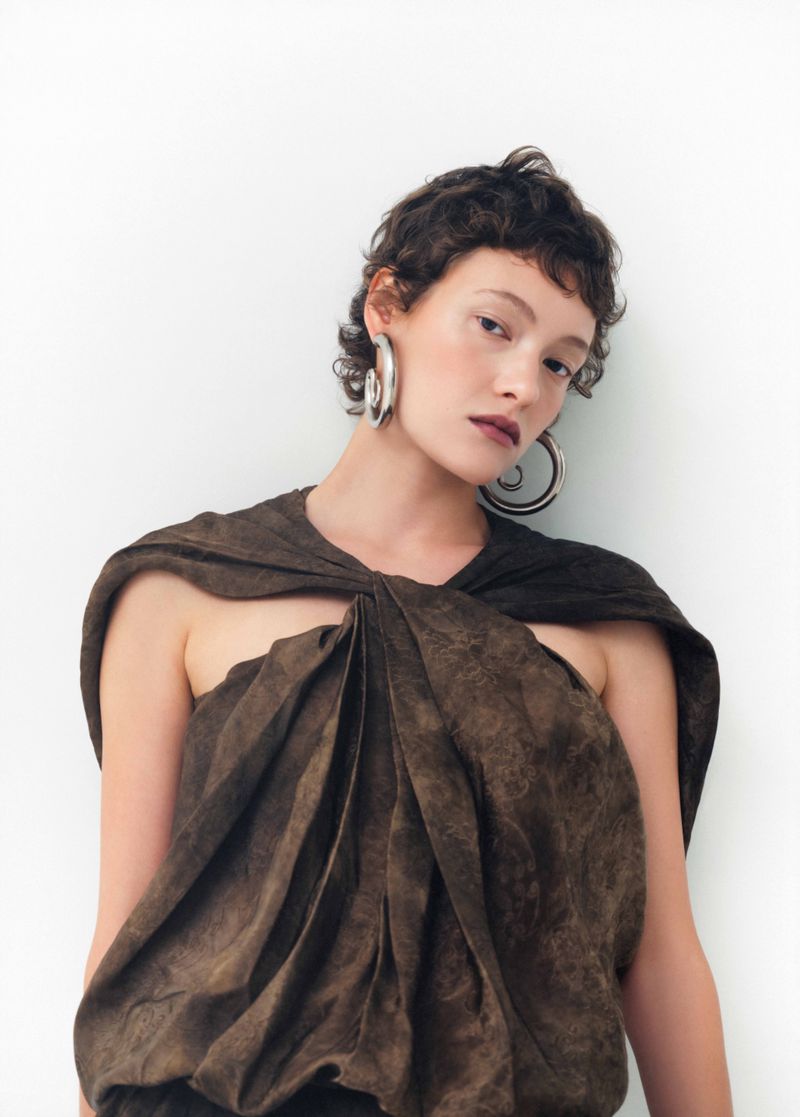 Model with dark short hair wearing oversized silver hoop earrings and a draped brown patterned top against a white background
