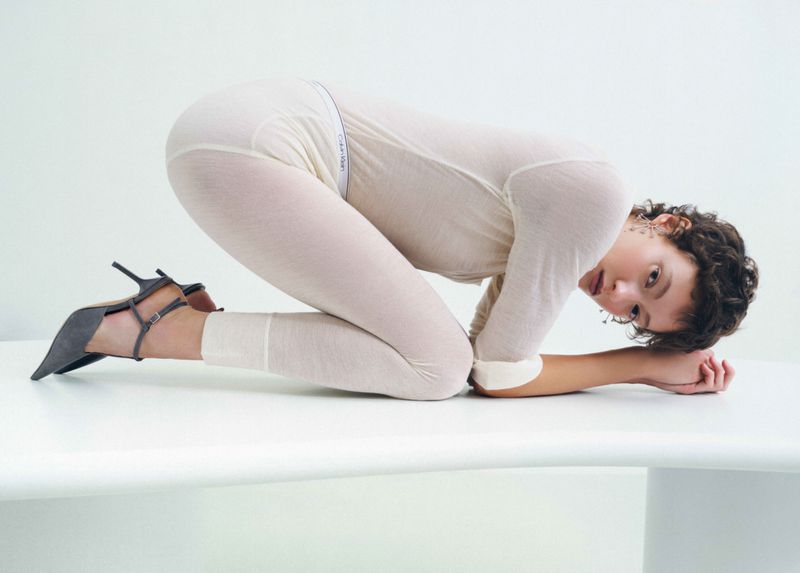 Model in cream bodysuit and dark heels kneeling on a white surface for an editorial project by Eleonora Adani.