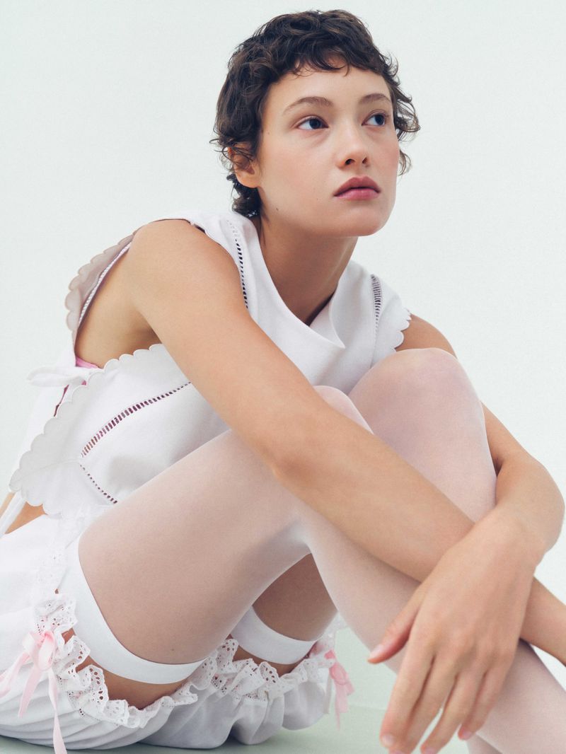 Model with short dark hair wearing a white sleeveless scalloped top and white stockings against a pale background