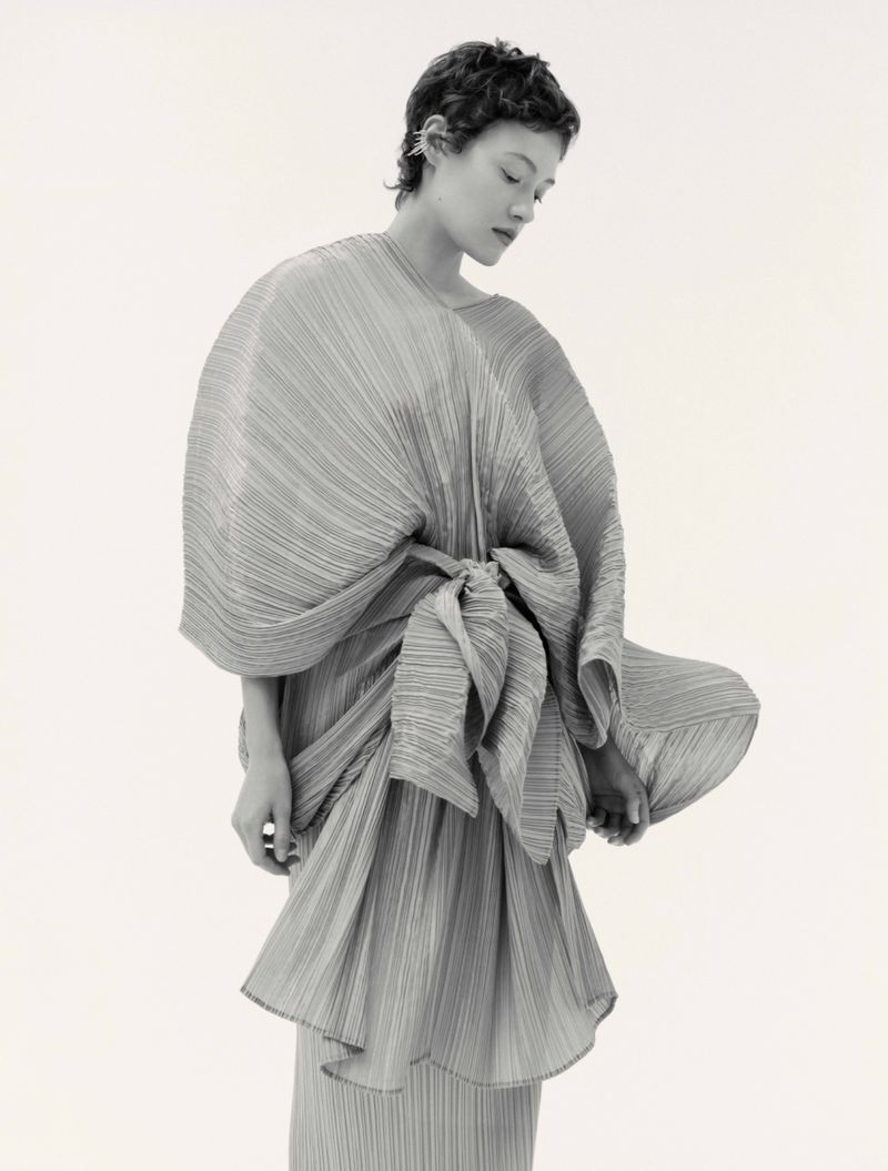 Model with short dark hair wearing a pleated draped dress against a plain white background