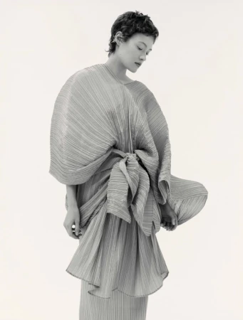 Model with short dark hair wearing a pleated draped dress against a plain white background