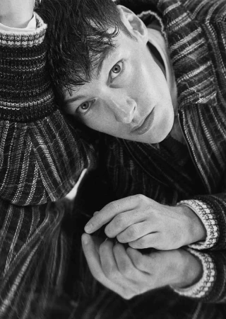 Close-up black and white portrait of a man wearing a patterned knit jacket, resting his head on his arm.