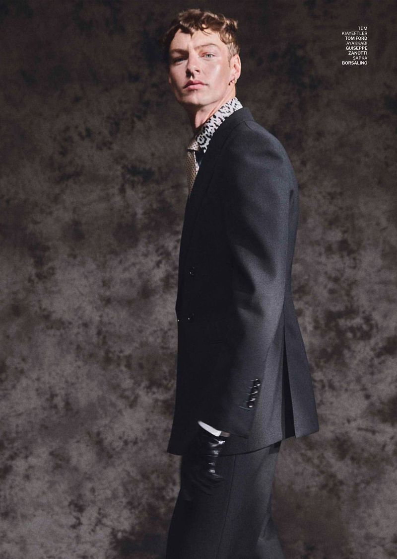 Model in a black suit jacket and dark trousers standing against a textured studio background