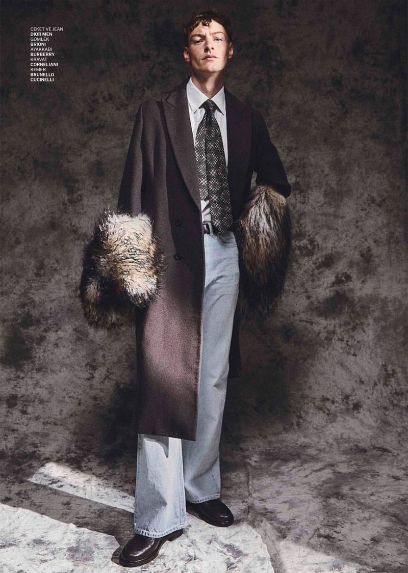 Male model wearing a dark coat with fur cuffs, light blue jeans, and a patterned tie against a mottled grey background.