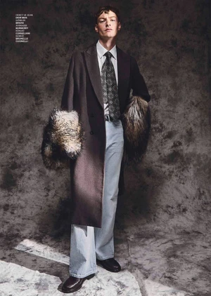 Male model wearing a dark coat with fur cuffs, light blue jeans, and a patterned tie against a mottled grey background.