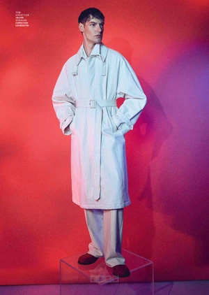 Full-length portrait of a male model wearing an off-white trench coat and trousers standing on a clear pedestal against a red backdrop