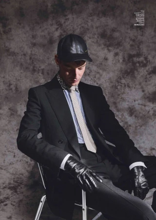 Man wearing a black suit, leather cap, and leather gloves seated against a textured grey studio backdrop.