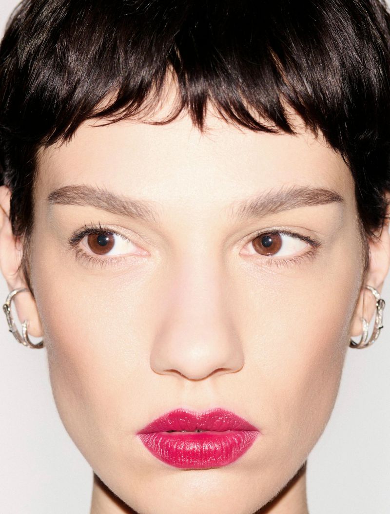 Close-up portrait of a woman with short dark hair, bold magenta lipstick, and silver curved earrings against a pale background.