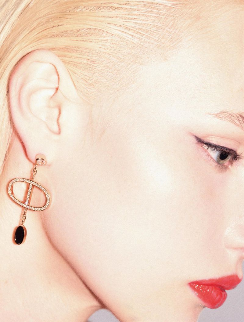 Close-up on fair-skinned woman's profile with red lipstick and blond hair, showcasing an elongated gold statement earring with a dark stone.