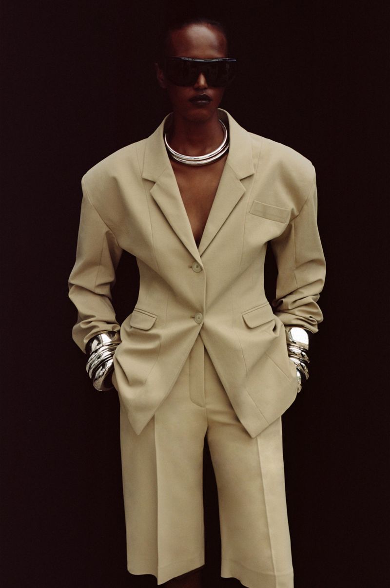 Model wearing a beige tailored blazer and shorts with dark sunglasses against a dark background