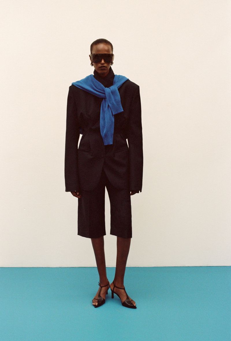 Model wearing dark blazer, shorts, sunglasses, and a blue sweater tied around the neck against a white and turquoise background.