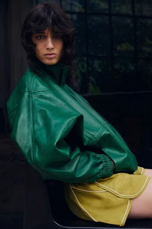 Model wearing a vibrant green leather jacket and shorts against a dark interior window background