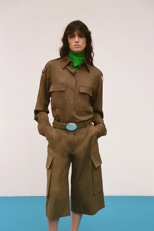 Model wearing a brown utility shirt and matching cargo shorts with a bright green turtleneck against a white and blue backdrop