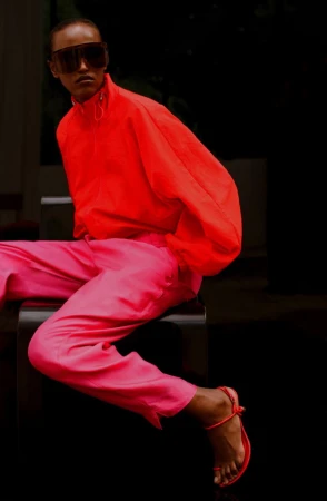 Model wearing a vibrant red jacket and pink trousers seated on a dark stool in a lookbook image for The Frankie Shop.