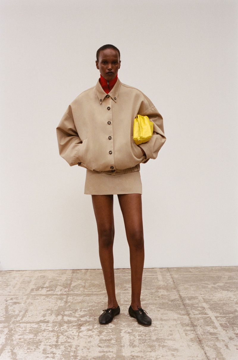 Model wearing an oversized beige jacket and matching skirt holding a yellow clutch against a plain wall.