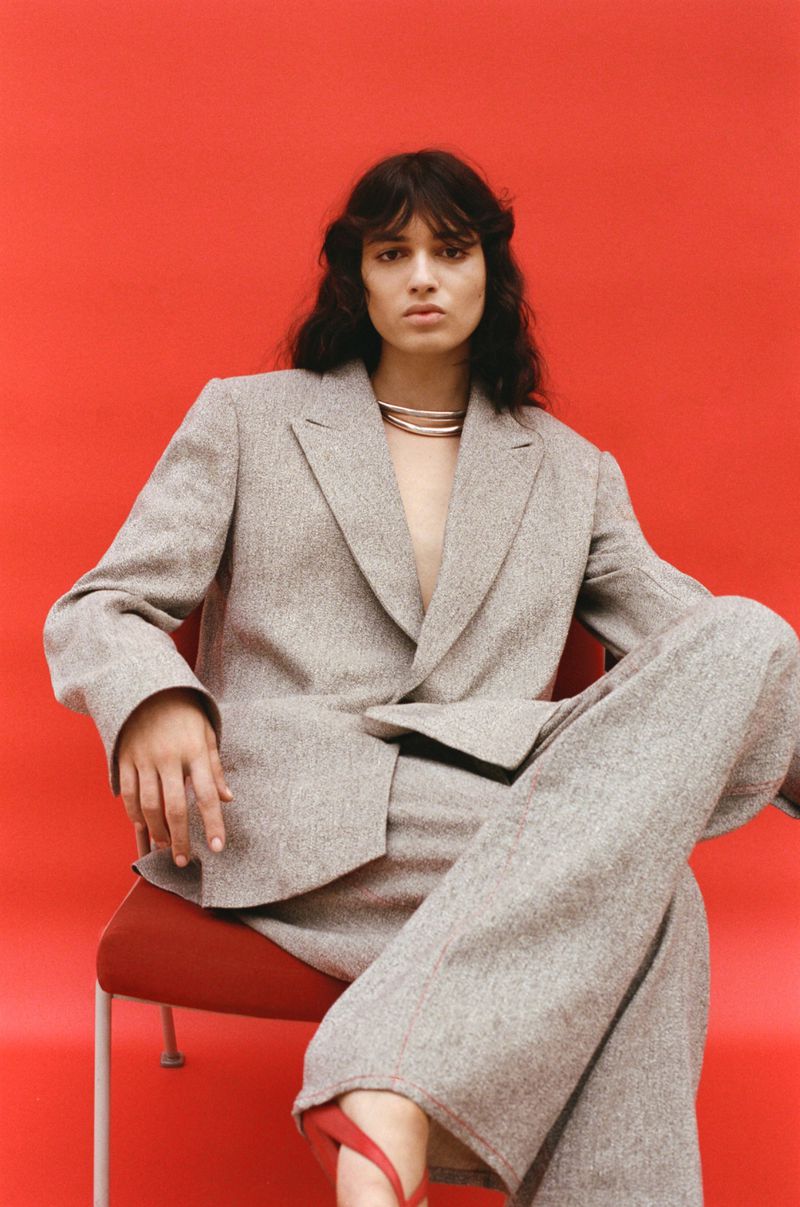 Model wearing a grey oversized blazer and matching trousers seated on a red chair against a vibrant red backdrop