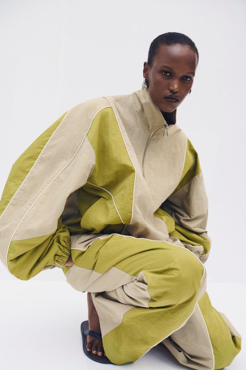 Model in olive green and beige panelled tracksuit crouching against a white background