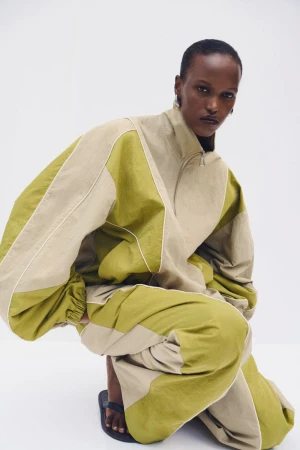 Model in olive green and beige panelled tracksuit crouching against a white background