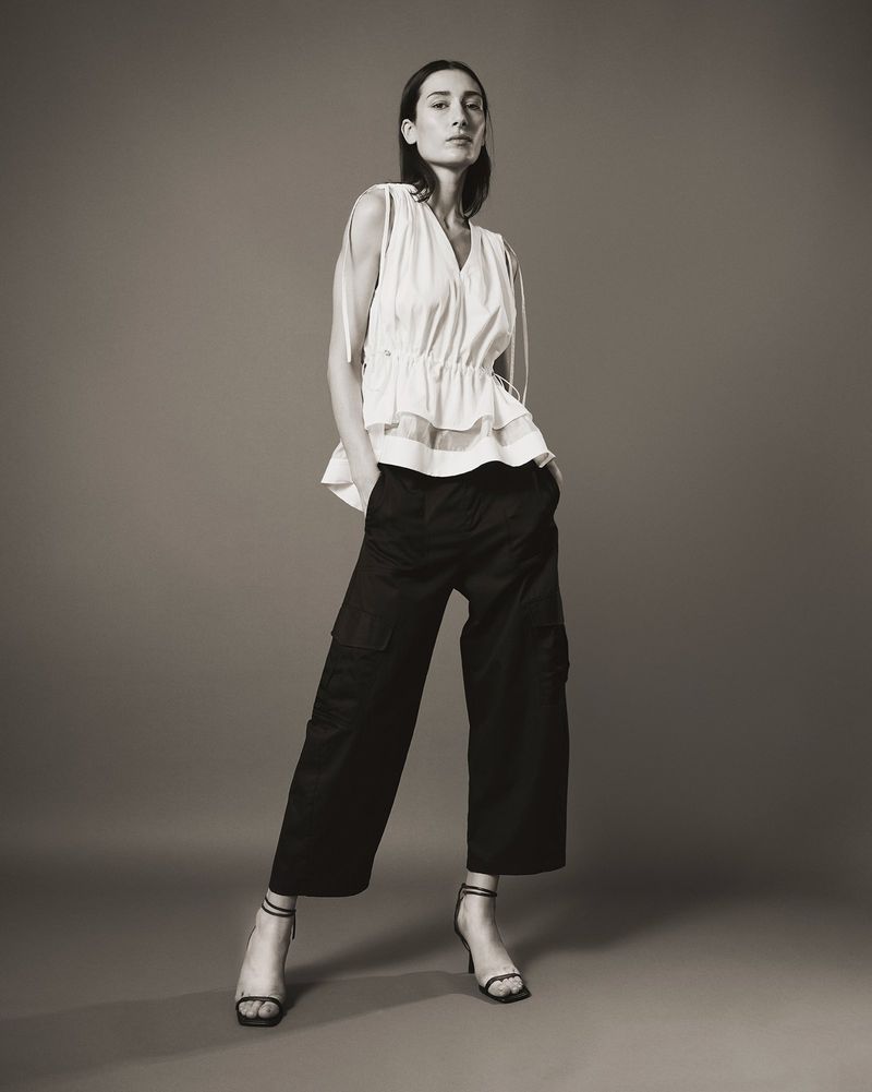 Model standing against a neutral studio backdrop wearing a white sleeveless top and dark trousers.