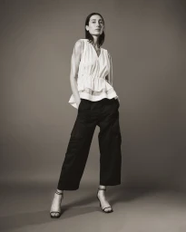 Model standing against a neutral studio backdrop wearing a white sleeveless top and dark trousers.