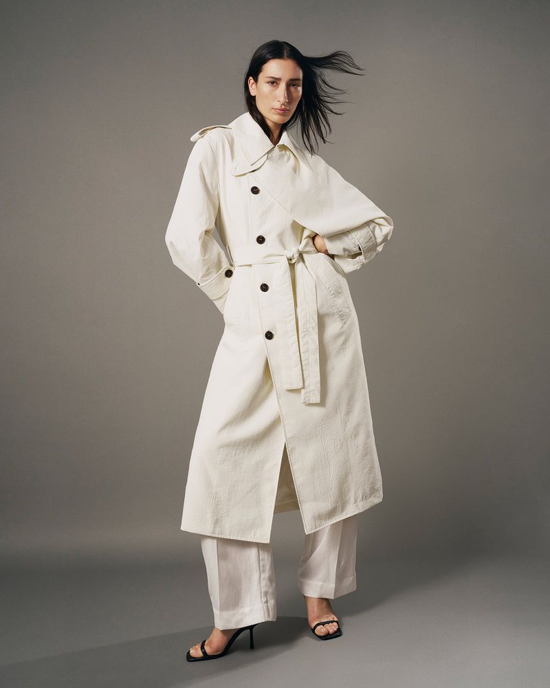 Model wearing a long white belted trench coat and matching trousers against a solid grey studio background.