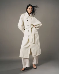 Model wearing a long white belted trench coat and matching trousers against a solid grey studio background.