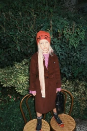 Child model standing in front of dense green foliage wearing an orange beanie, brown coat, and black handbag