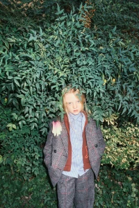 Child model wearing a tailored plaid suit and striped shirt framed by dense green foliage in a fashion lookbook image.