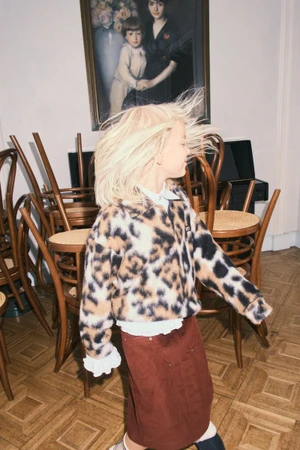 Child with blonde hair in motion wearing a leopard print jacket and brown skirt inside a room with wooden chairs