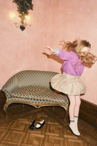 A young girl with long blond hair jumping in mid-air in front of a patterned vintage sofa and pink wall.
