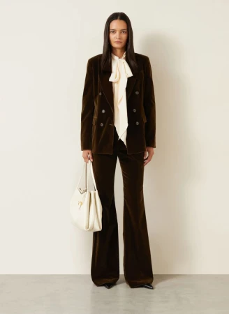 Model wearing a dark brown velvet suit with cream blouse and white handbag against a plain background