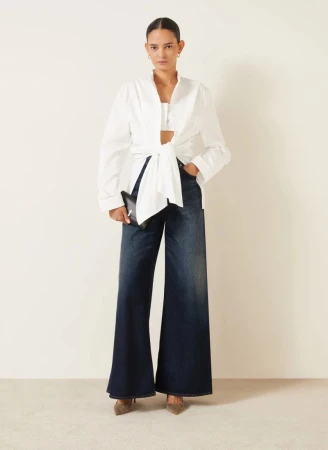 Model Wanessa Milhomem wearing a white tied shirt and wide leg denim jeans holding a black clutch against a plain background