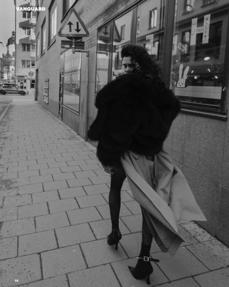 Monochrome fashion shot of a woman walking on a city sidewalk wearing a fur coat, stockings, heels, and a trench coat over her shoulder.