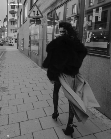Monochrome fashion shot of a woman walking on a city sidewalk wearing a fur coat, stockings, heels, and a trench coat over her shoulder.