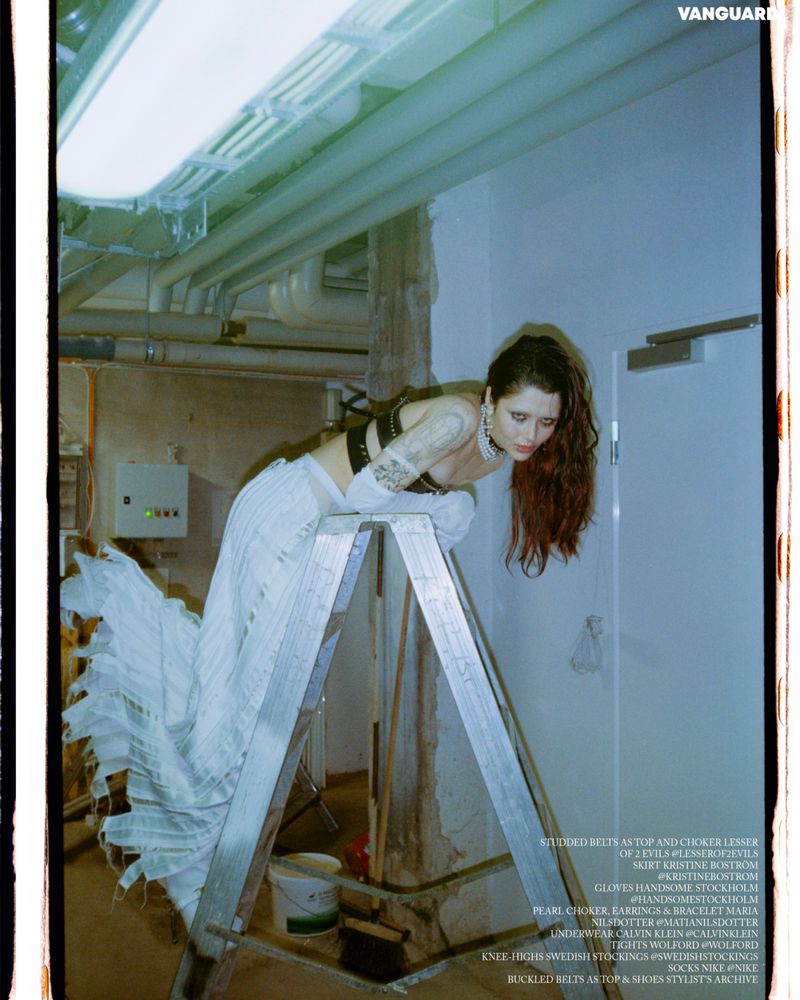 A woman with auburn hair poses on a ladder in an unfinished room wearing a studded crop top and skirt for a fashion editorial.