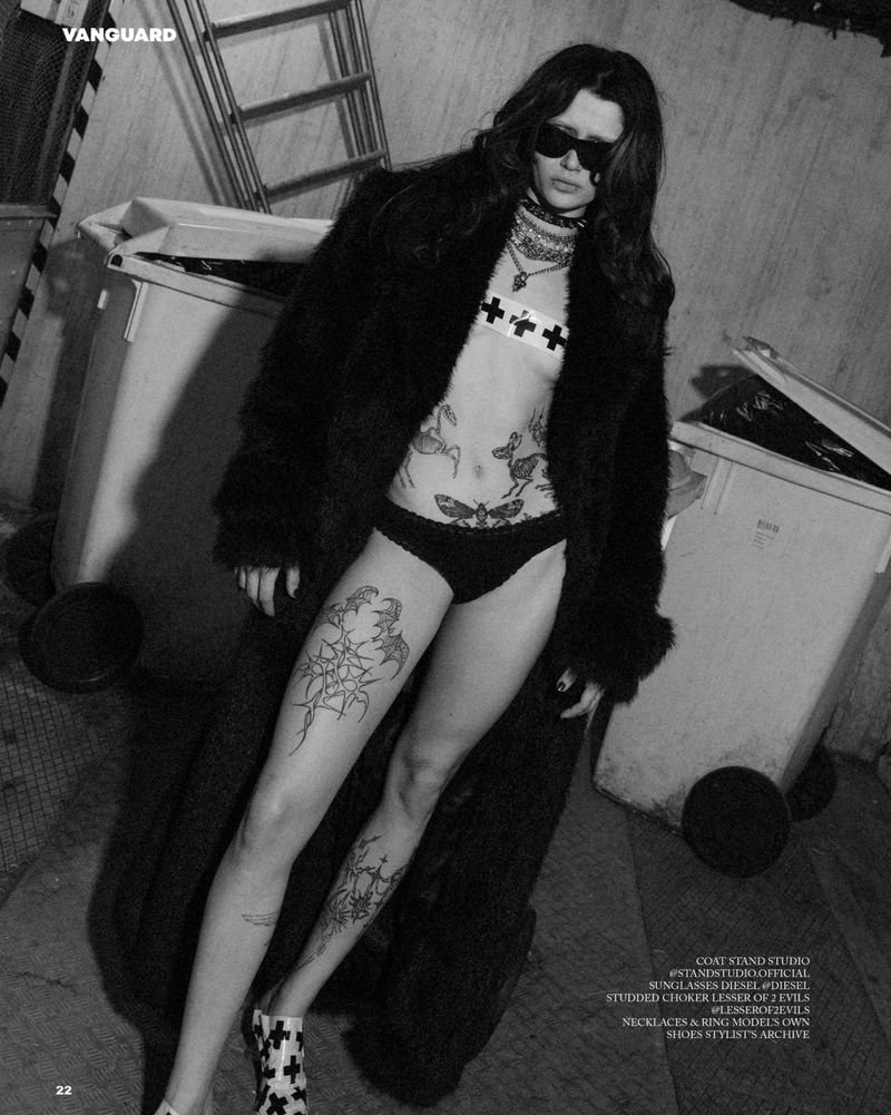Black-and-white fashion shot: woman in sunglasses, lingerie, and fur coat poses near industrial bins with visible tattoos and bold footwear.