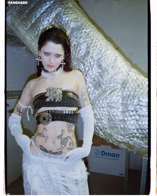 Edgy fashion photo: Woman with dramatic eye makeup, black leather belts, and tattoos poses indoors with a metallic pipe behind her.