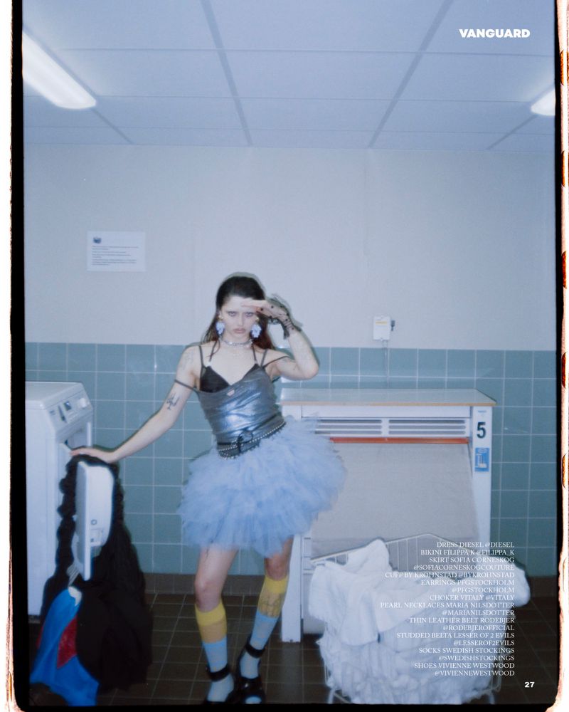 A young woman poses in a laundry room wearing a silver top, blue tutu, and striped socks, holding her hand up to her forehead.