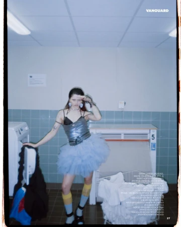 A young woman poses in a laundry room wearing a silver top, blue tutu, and striped socks, holding her hand up to her forehead.