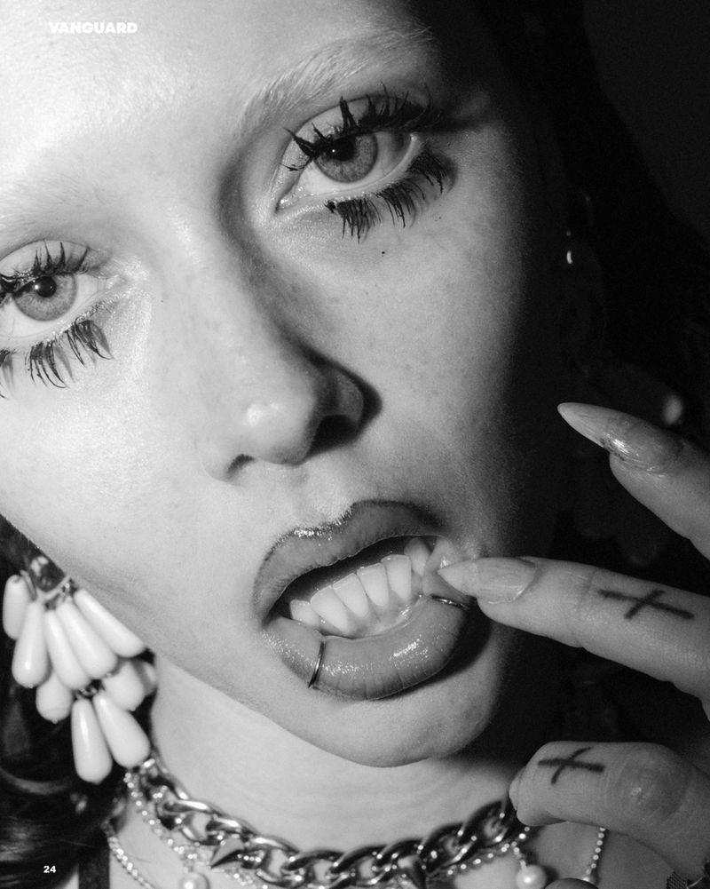 Close-up black and white fashion shot of a woman's face with striking makeup, jewelry, and distinctive pierced lip and finger tattoos.