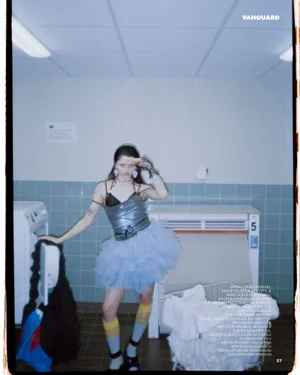 Fashion model in a silver top and blue tulle tutu skirt posing beside a washing machine in a laundromat setting.