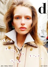Close-up portrait of a model with light brown hair wearing a collared shirt and beige coat in an urban street setting