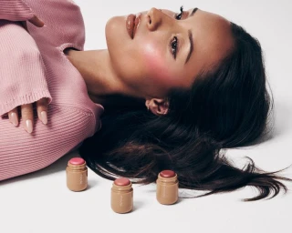 Woman in a pink knitted top lying down next to three small cosmetic jars against a clean white background