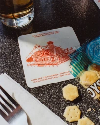 Custom drink coaster with red illustration of Ray's Waterfront set on a dark granite table next to crackers and a glass.