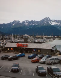 Elevated view of a waterfront restaurant with parked cars in the foreground and snowy mountains behind a marina.